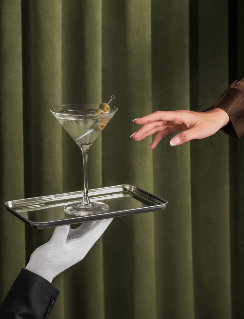 A gloved hand offers a martini on a silver tray as another hand reaches for it